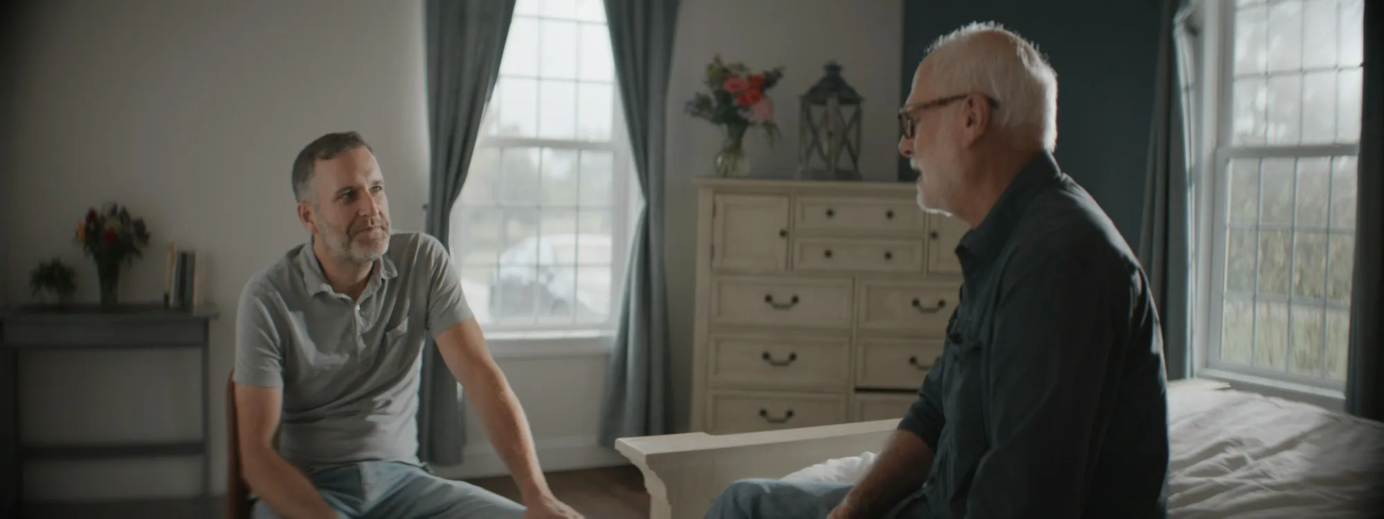 An older man talking with a visitor in a sunlit bedroom